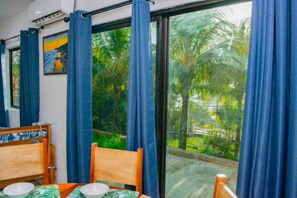 Fondo de ventanas con cortinas azules y vista a palmeras tropicales desde interior de alojamiento.