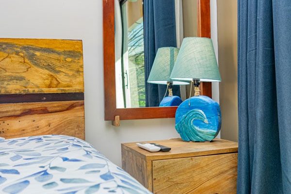 Reflejo en espejo de madera, cama con ropa de cama azul y blanca, lámpara con tema de olas, cortinas azules, ambiente acogedor en habitación de surf.